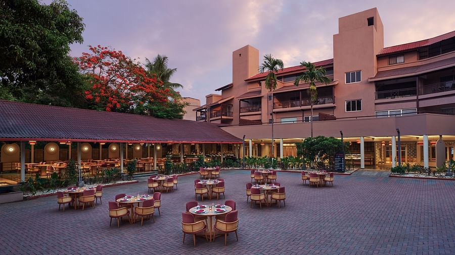Outdoor dining setup at Courtyard Sky Restro at DGV Resort by Treat, Silvassa with round tables and chairs at a resort, surrounded by trees and multi-storey buildings at dusk