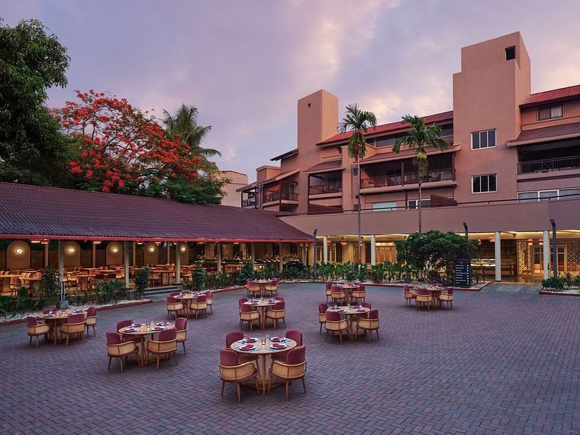 Outdoor dining setup at Courtyard Sky Restro at DGV Resort by Treat, Silvassa with round tables and chairs at a resort, surrounded by trees and multi-storey buildings at dusk