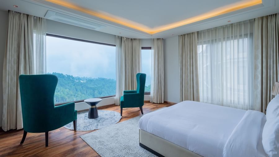 Spacious, sunlit suite with floor-to-ceiling windows revealing breathtaking mountain views, a modern white bed, and a turquoise armchair, highlighting luxurious accommodations at The Manor Sports & Wellness Hotel in Shimla.