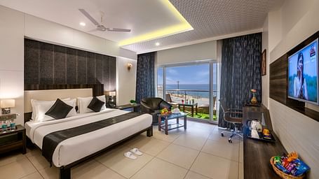 A bright room with a king size bed, seating area, workspace, TV, and balcony overlooking a view.