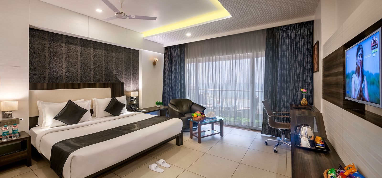 A modern hotel room featuring a king sized bed with black and white accents, a seating area, a work desk, and a wall-mounted TV.