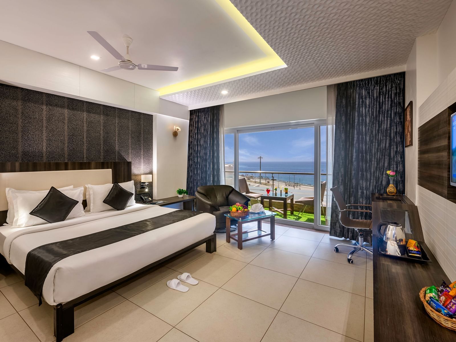 A bright room with a king size bed, seating area, workspace, TV, and balcony overlooking a view.
