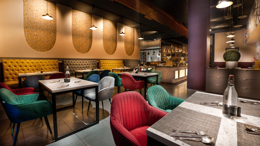 An inviting dining area featuring plush red, green, and blue chairs at neatly set wooden tables under warm decorative wall lighting at Apartel by Aarin, Oragadam.