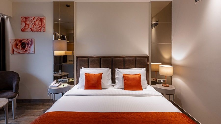 The bedroom features a bed with white linens and contarsting red pillows, with lamp shades on both the sides of the bed and a brown head rest,  has two paintings enhancing the wall space behind the bed.