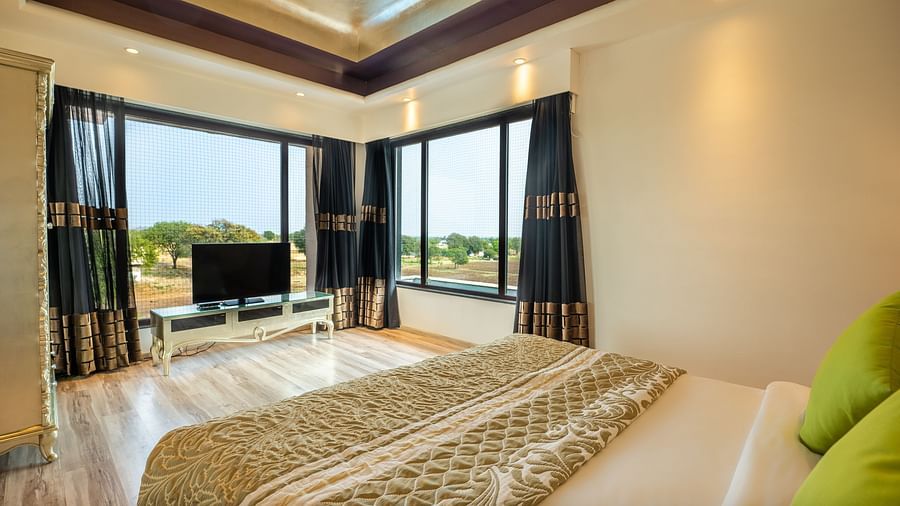The bed at Crescent Spa & Resort, Indore, is spacious with a photo frame above the headboard, two warmly lit bedside tables, and two large windows with curtains pulled to the side