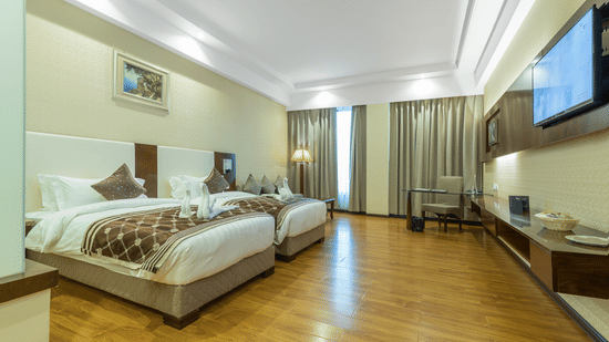 Modern hotel bedroom with twin beds, patterned runners, a long wooden desk with a wall-mounted TV, and polished wooden flooring at Golden Tulip Kukas, Jaipur.