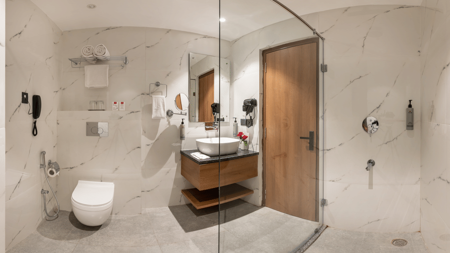 A tiled bathroom at Lords Inn Katra showing a glass shower partition, a toilet, a vanity counter with a mirror, and a washbasin.