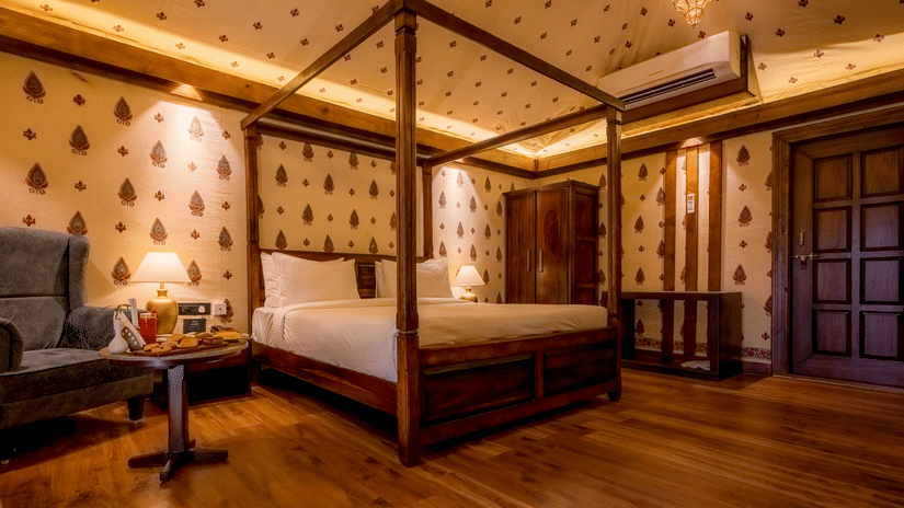 A Premium Tent accommodation space featuring poster bed, a sofa chair, a centre table with snacks and beverages, bedside table on either side with lamps, a wooden wardrobe, AC, and lot more at The Baagh Ananta Elite, Ranthambore