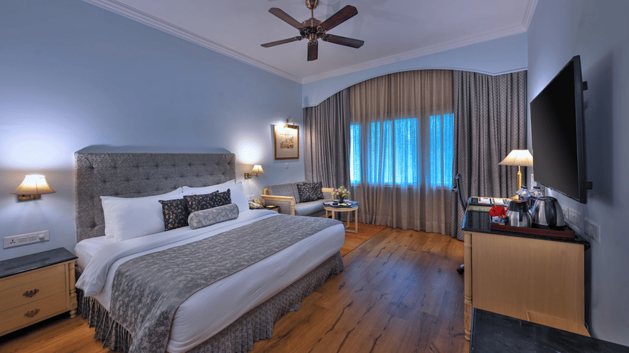 A room with a large bed, spreads, pillows, a fan, and windows at The Clarks Varanasi.
