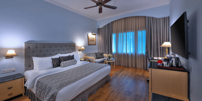 A room with a large bed, spreads, pillows, a fan, and windows at The Clarks Varanasi.