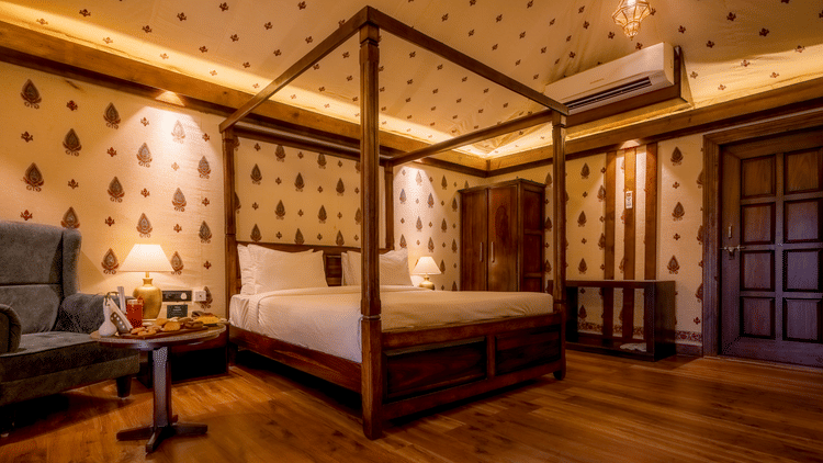 A Premium Tent accommodation space featuring poster bed, a sofa chair, a centre table with snacks and beverages, bedside table on either side with lamps, a wooden wardrobe, AC, and lot more at The Baagh Ananta Elite, Ranthambore