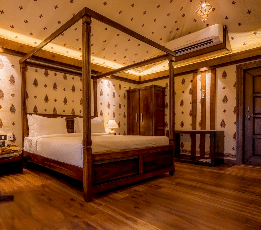A Premium Tent accommodation space featuring poster bed, a sofa chair, a centre table with snacks and beverages, bedside table on either side with lamps, a wooden wardrobe, AC, and lot more at The Baagh Ananta Elite, Ranthambore