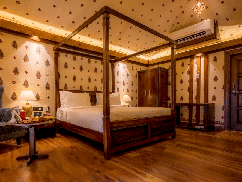 A Premium Tent accommodation space featuring poster bed, a sofa chair, a centre table with snacks and beverages, bedside table on either side with lamps, a wooden wardrobe, AC, and lot more at The Baagh Ananta Elite, Ranthambore