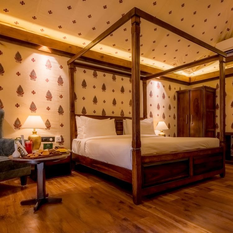 A Premium Tent accommodation space featuring poster bed, a sofa chair, a centre table with snacks and beverages, bedside table on either side with lamps, a wooden wardrobe, AC, and lot more at The Baagh Ananta Elite, Ranthambore