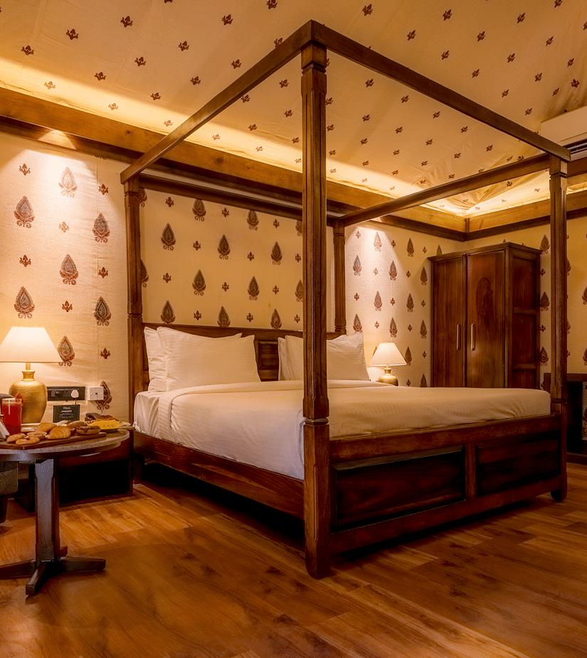 A Premium Tent accommodation space featuring poster bed, a sofa chair, a centre table with snacks and beverages, bedside table on either side with lamps, a wooden wardrobe, AC, and lot more at The Baagh Ananta Elite, Ranthambore