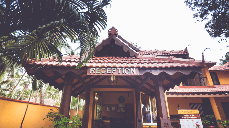 Reception entrance with red-tiled roof, wooden doors, and signage, surrounded by palm trees at Paradise Lagoon Resort, Udupi.