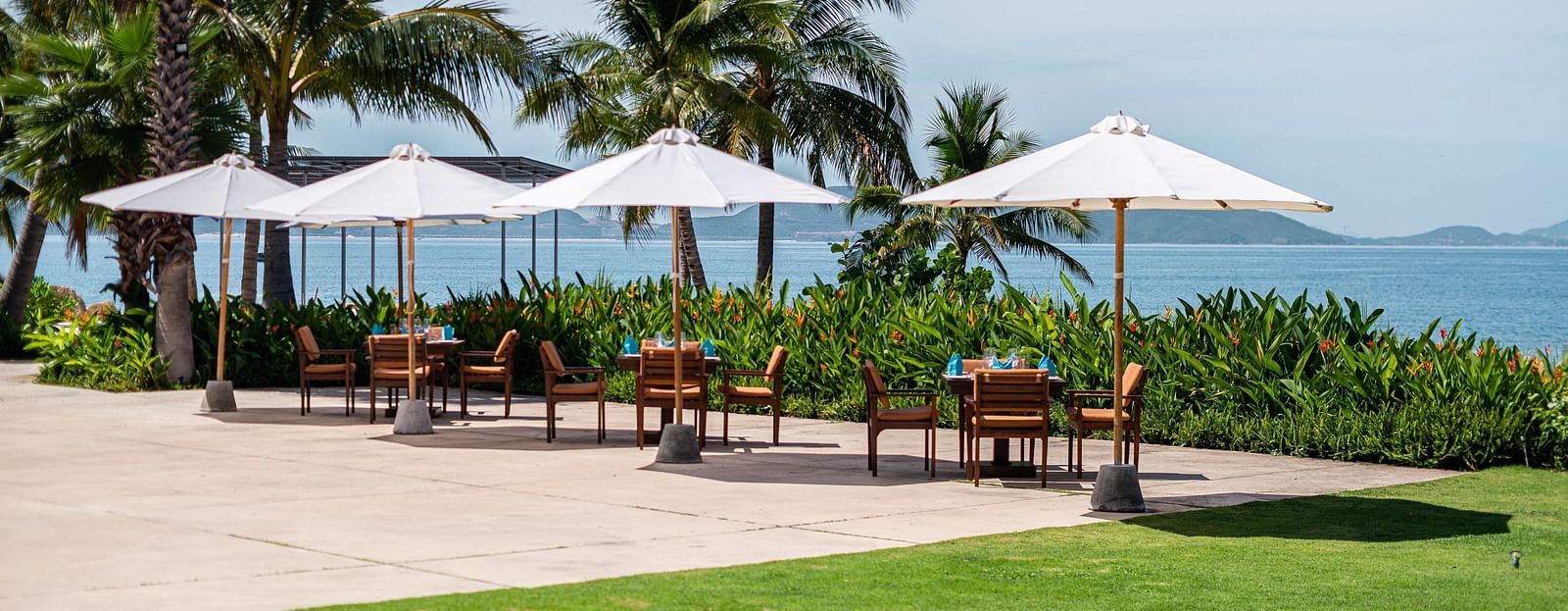 A tropical outdoor restaurant scene with white parasols, palm trees and a view of the ocean and distant islands - Alibu Resorts Nha Trang