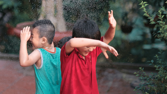 Two children laughing and playing in water sprinklers outdoors, shielding themselves from the spray on a sunny day.