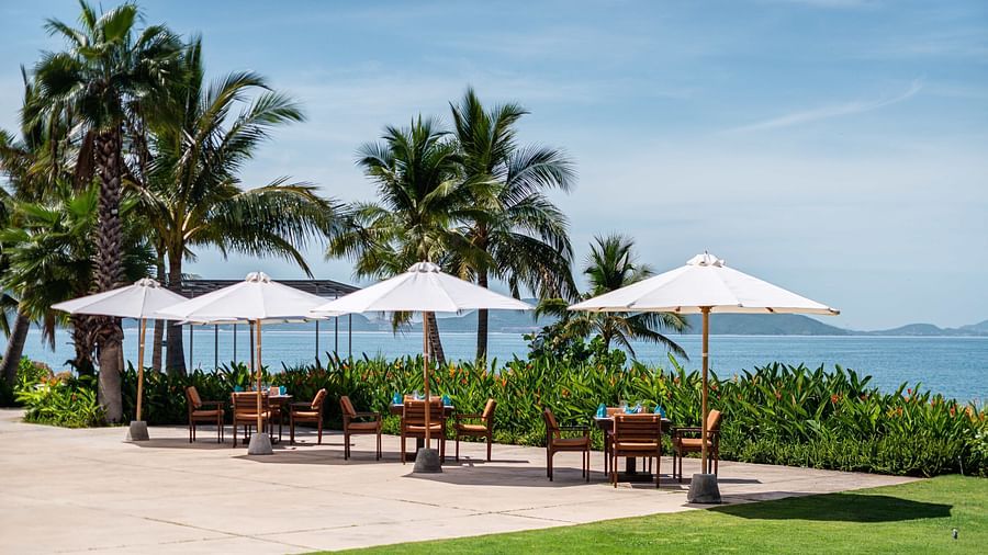 A tropical outdoor restaurant scene with white parasols, palm trees and a view of the ocean and distant islands - Alibu Resorts Nha Trang