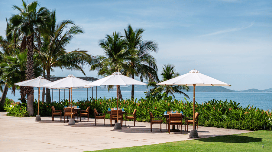 A tropical outdoor restaurant scene with white parasols, palm trees and a view of the ocean and distant islands at Alibu Resorts Nha Trang