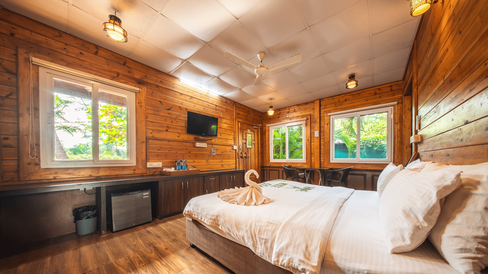 Head-corner view of a cozy room at Tranquil Beach Resort, Harihareshwar, featuring wooden walls, a neatly made bed, a small table, and windows bringing in natural light.