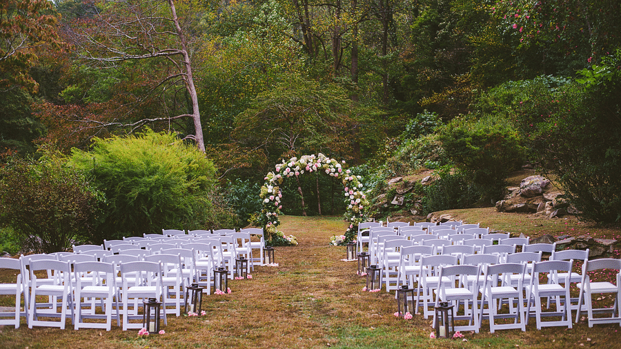 a beautiful setup for a wedding with white colored folding chairs  featuring an arch covered with flowers in the middle of the aisle - Historic Tapoco Lodge