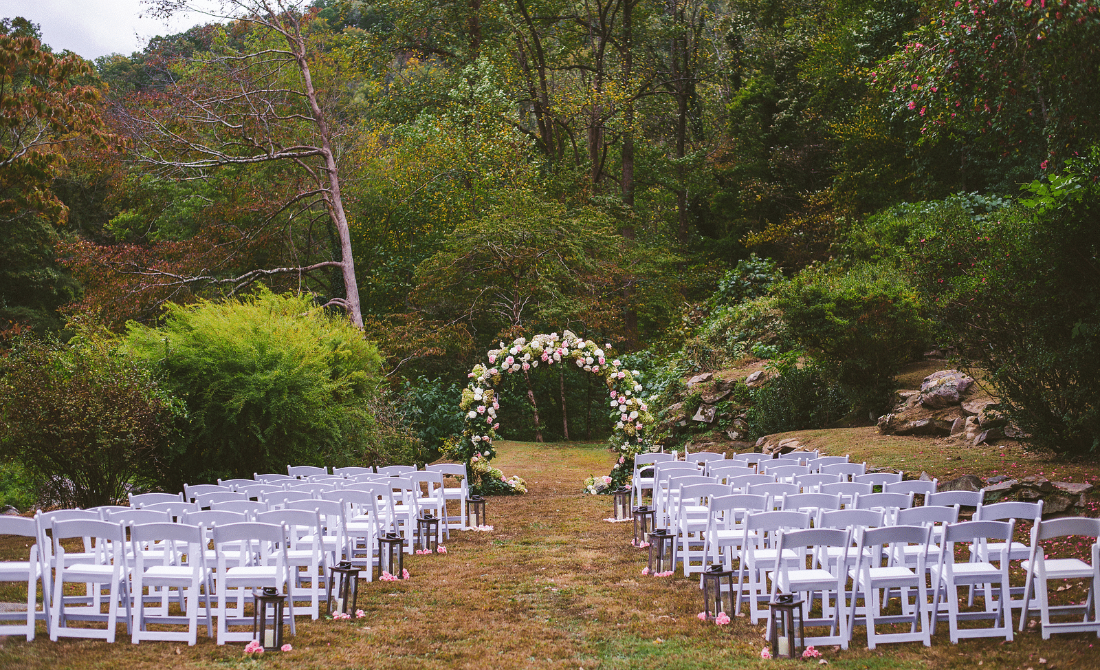 a beautiful setup for a wedding with white colored folding chairs  featuring an arch covered with flowers in the middle of the aisle - Historic Tapoco Lodge