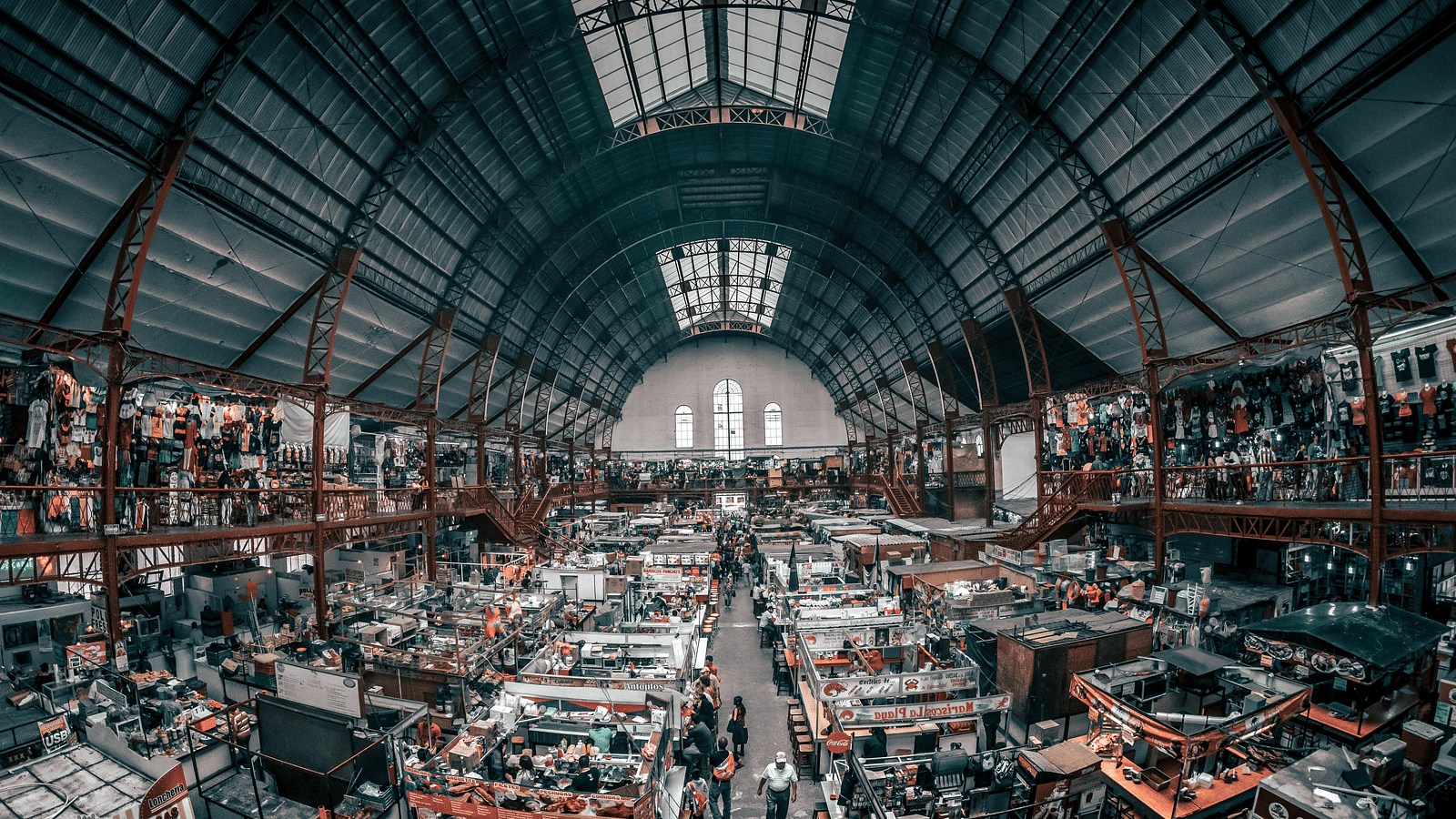 an aerial view of a market with numerous stalls in two levels with people browsing what they want to buy