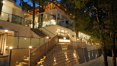 Multi-tiered hillside resort nestled among tall trees, featuring illuminated stone staircases, glass railings, and elevated terraces at DLS The Emerald, Nainital.