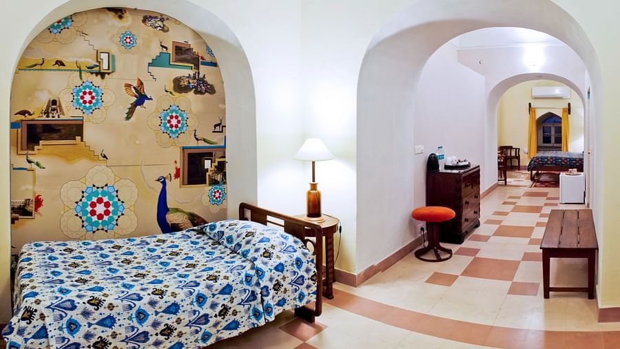 Hotel room with a single bed featuring blue patterned bedding, a bedside table with a lamp in a decorated alcove, and a view through an archway to a hallway with furniture at Tijara Fort-Palace, Alwar.