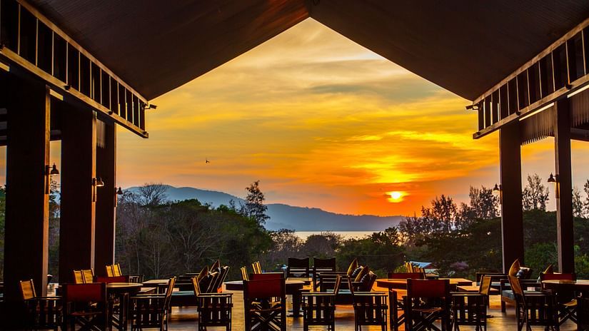 Wooden chairs in an open restaurant that overlooks a setting sun and mountains - Symphony Samudra Beachside Jungle Resort And Spa, Port Blair