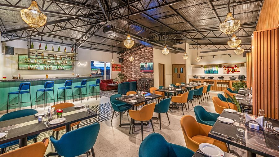 A restaurant with colourful chairs, dark tables, and a long bar with blue stools. It features a high ceiling with exposed beams, hanging pendant lights, and a buffet area in the background - Lords Eco Inn Dehradun.