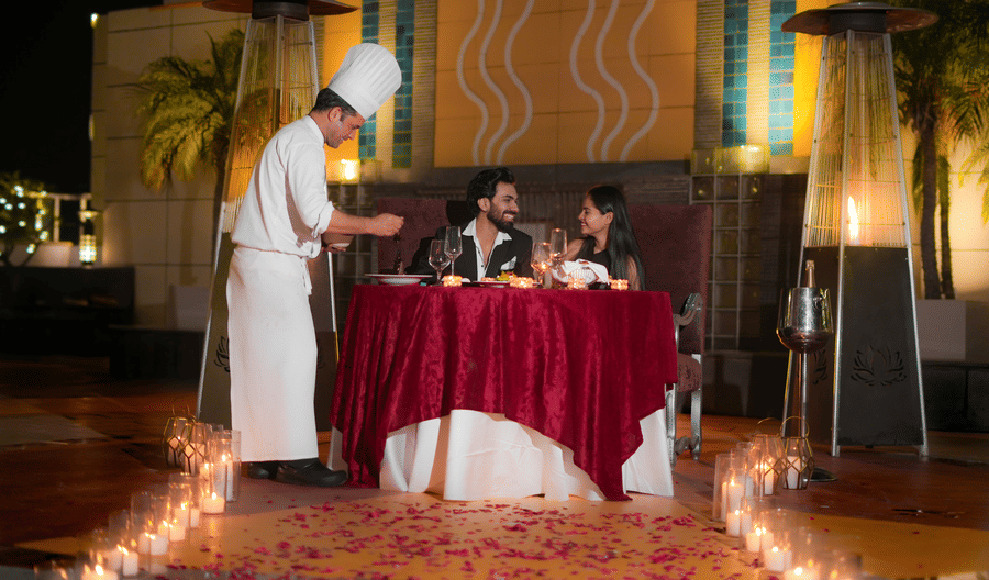 A chef standing beside a table serving food to a seated couple at an outdoor dining setup with candles at The Suryaa, New Delhi.