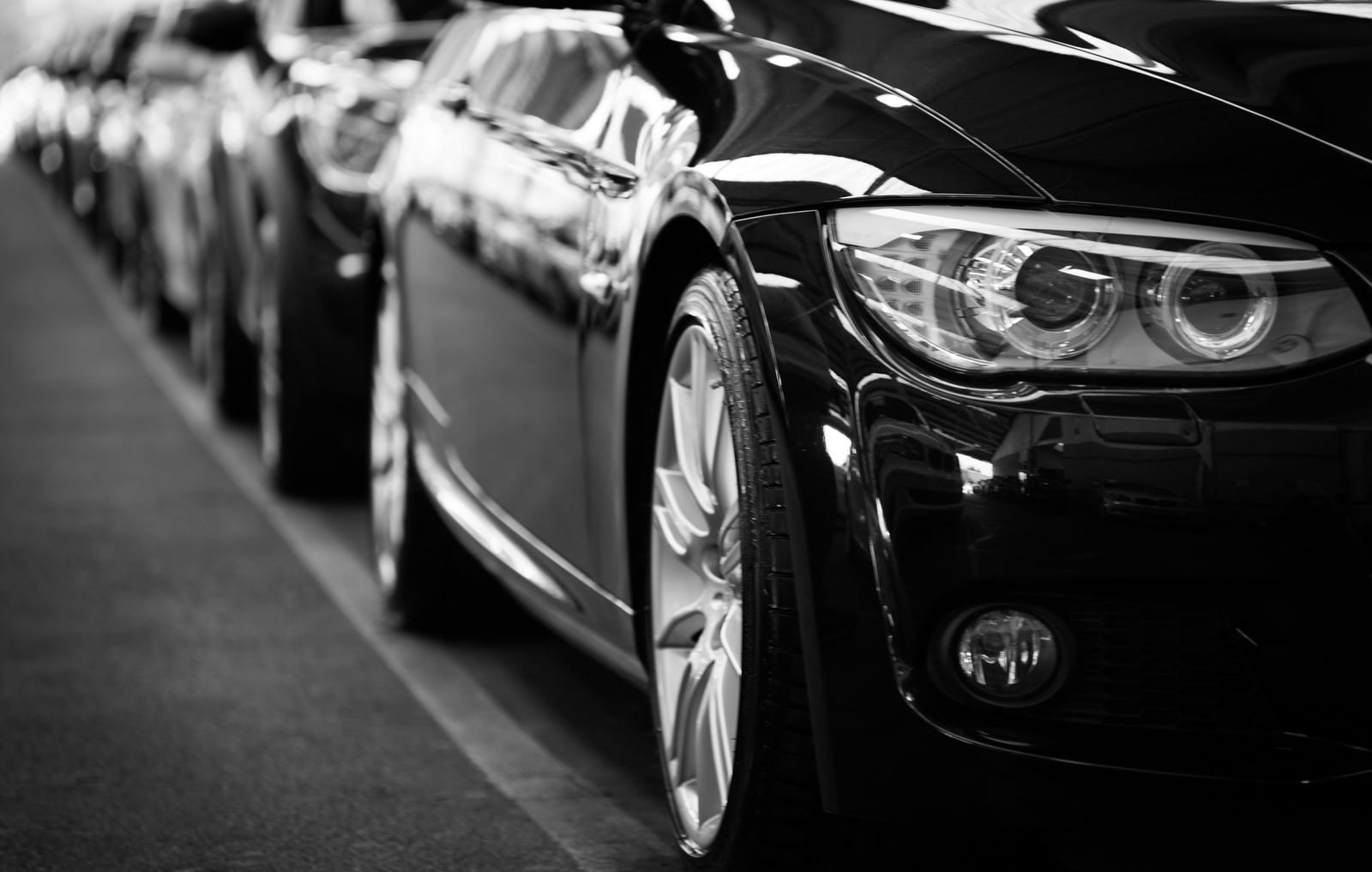 A close-up, black and white shot shows a line of sleek, dark-coloured cars parked closely together, emphasising their headlights and front wheels.