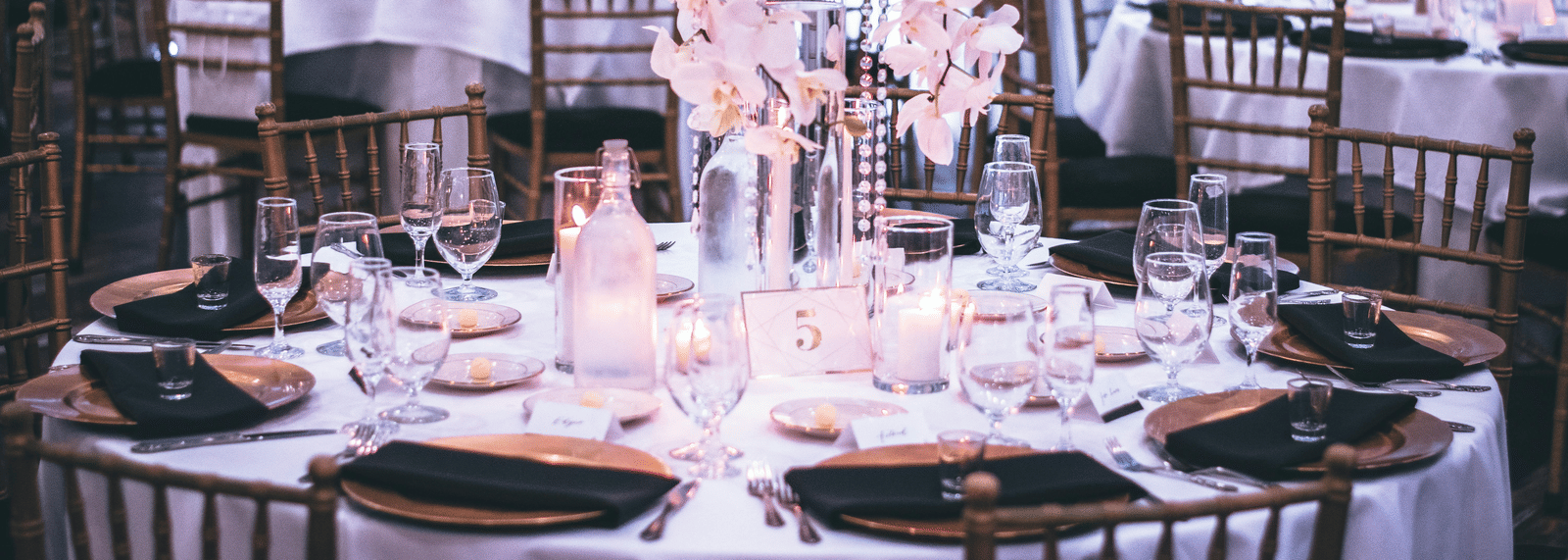 A round table with a white table cloth along with huge plates set on the table and a beautiful flower vase in the middle.
