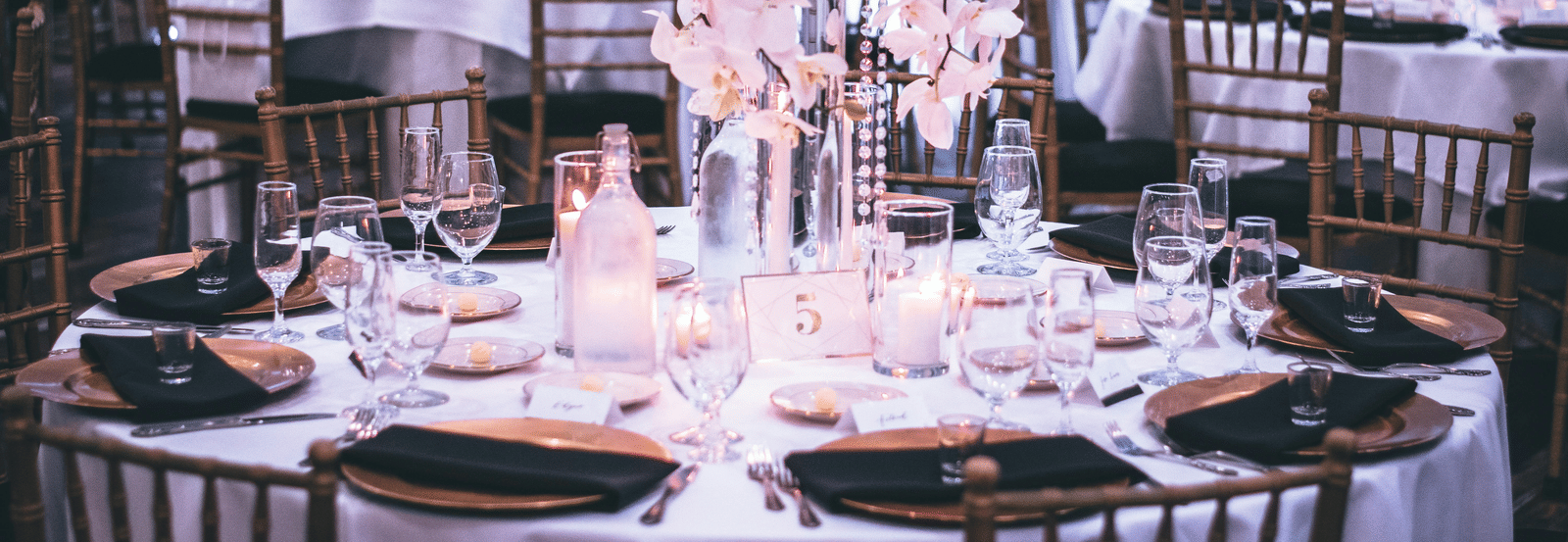 A round table with a white table cloth along with huge plates set on the table and a beautiful flower vase in the middle.