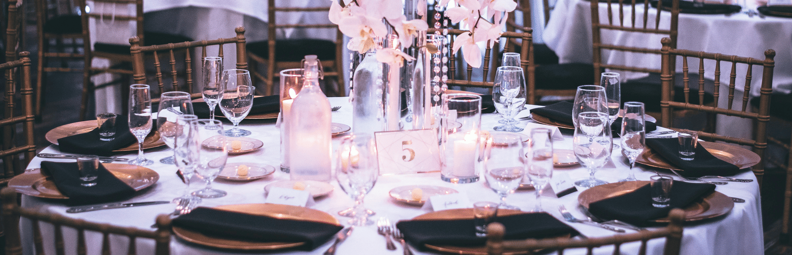A round table with a white table cloth along with huge plates set on the table and a beautiful flower vase in the middle.