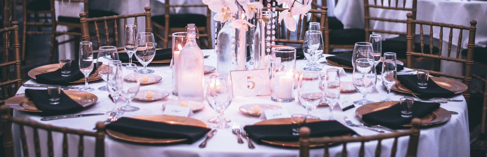 A round table with a white table cloth along with huge plates set on the table and a beautiful flower vase in the middle.