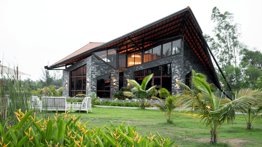 Exterior view of a modern stone and glass building with a sloping roof, green lawn, and tropical plants at Hotel Sonar Bangla, Sundarban.