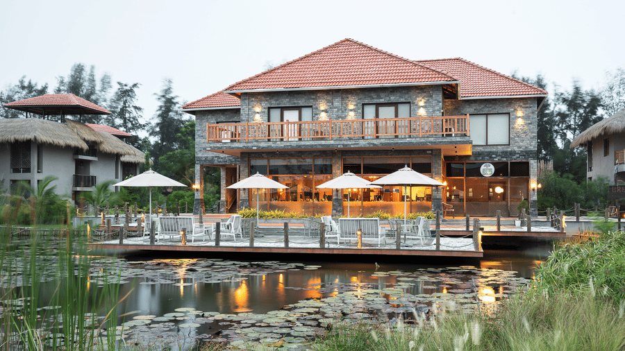Front view of a stone-clad building with a red tiled roof featuring a ground-floor restaurant area and a wooden terrace extending over a pond at Hotel Sonar Bangla, Sundarban.