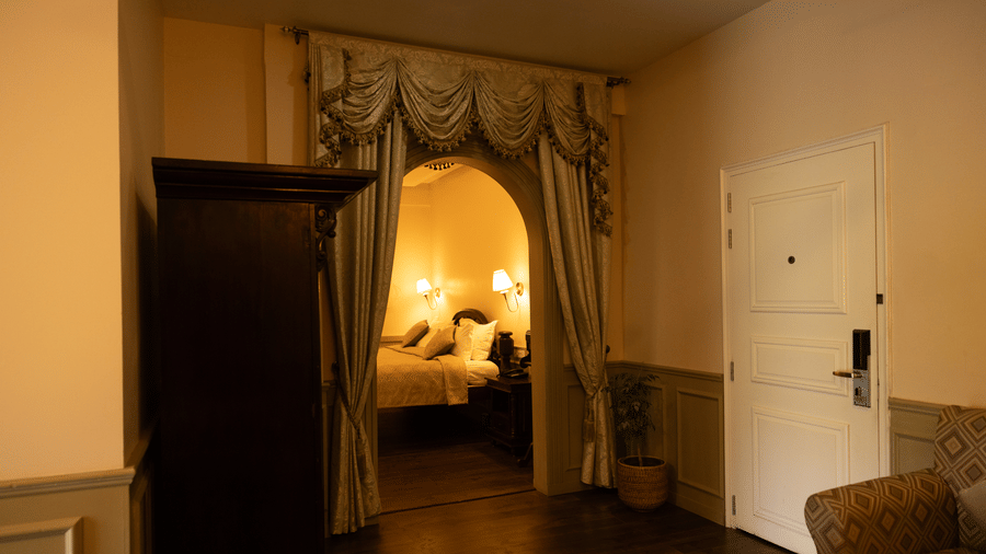 A room at The Heritage Club, Tripura, Castle, Shillong, showcasing a space with a wardobe and a door, leading to the bedroom with a large bed, pillows, and lamps.
