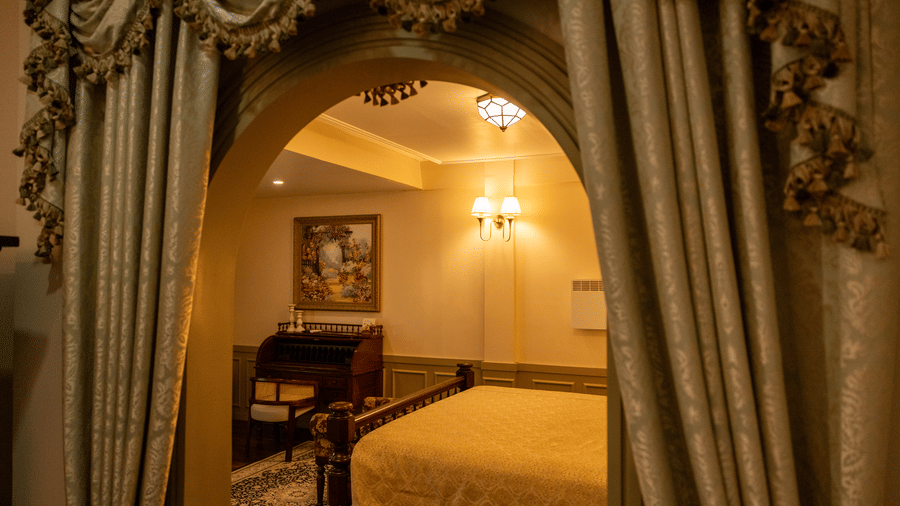 A room at The Heritage Club, Tripura, Castle, Shillong, showcasing a space leading to a bedroom with a large bed, spreads, wall art, and lamps.