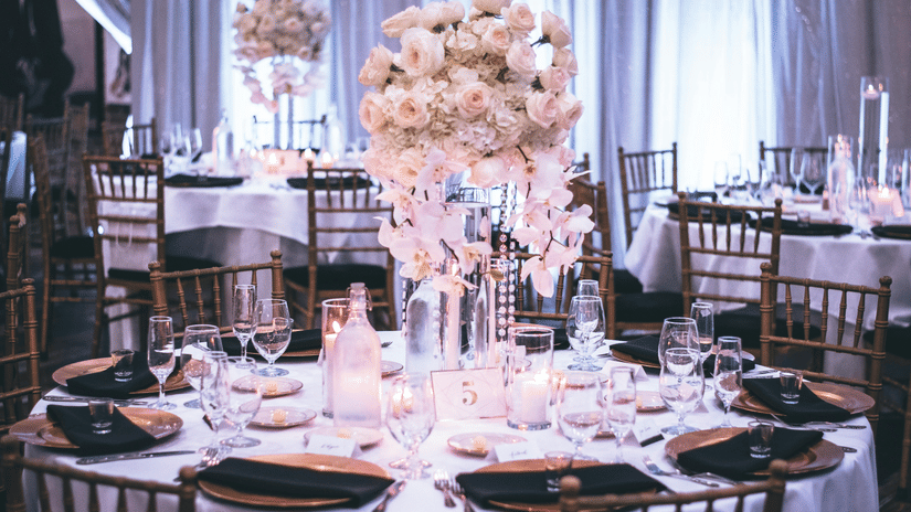 A round table with a white table cloth along with huge plates set on the table and a beautiful flower vase in the middle.