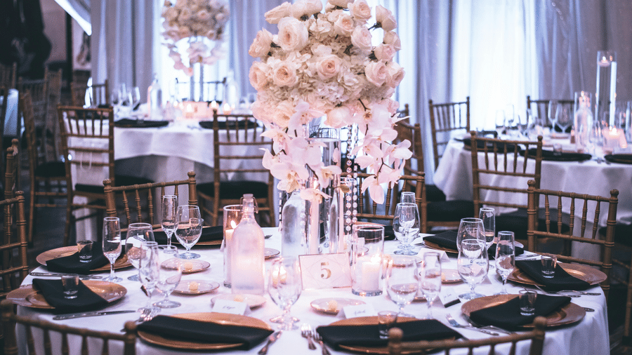 A round table with a white table cloth along with huge plates set on the table and a beautiful flower vase in the middle.