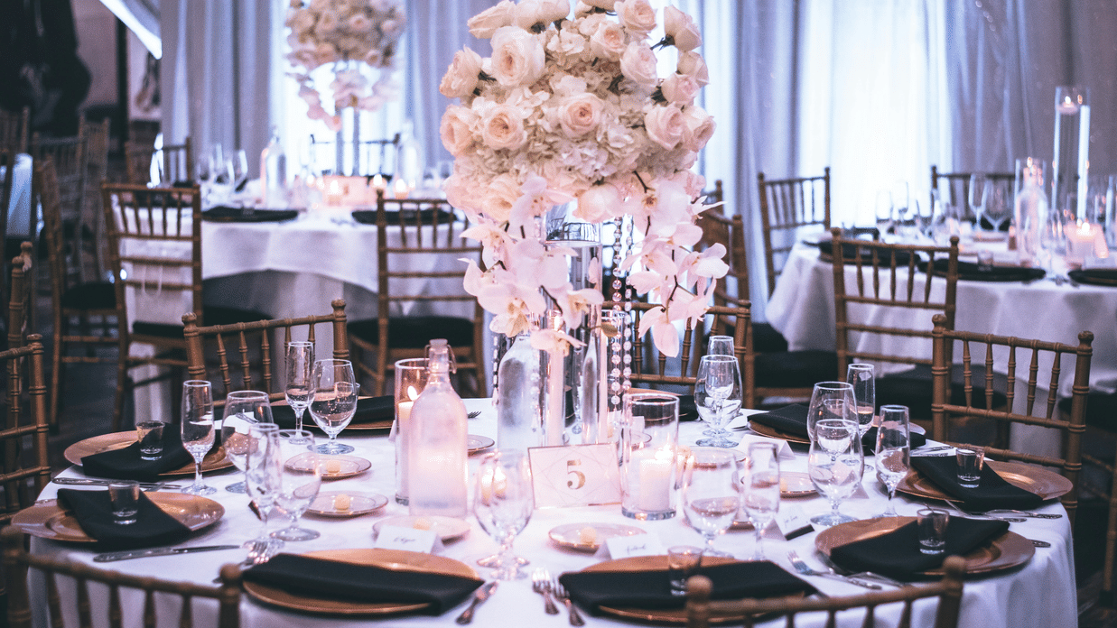 A round table with a white table cloth along with huge plates set on the table and a beautiful flower vase in the middle.