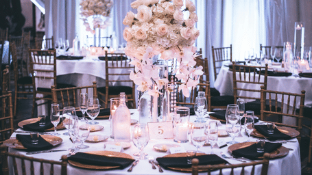 A round table with a white table cloth along with huge plates set on the table and a beautiful flower vase in the middle.