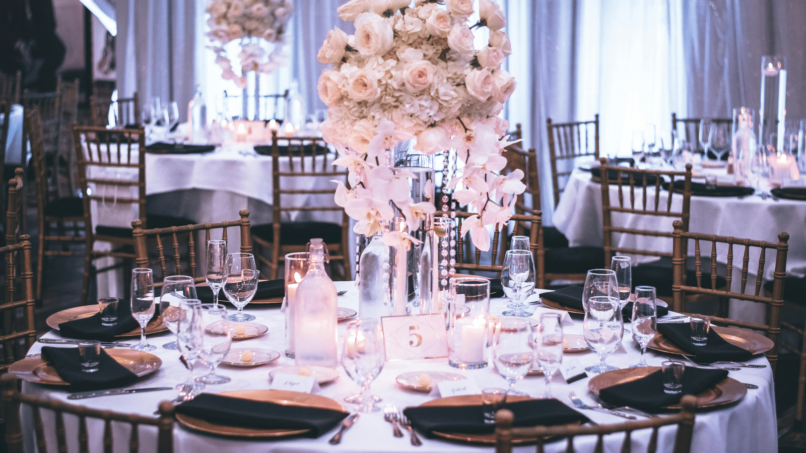 A round table with a white table cloth along with huge plates set on the table and a beautiful flower vase in the middle.