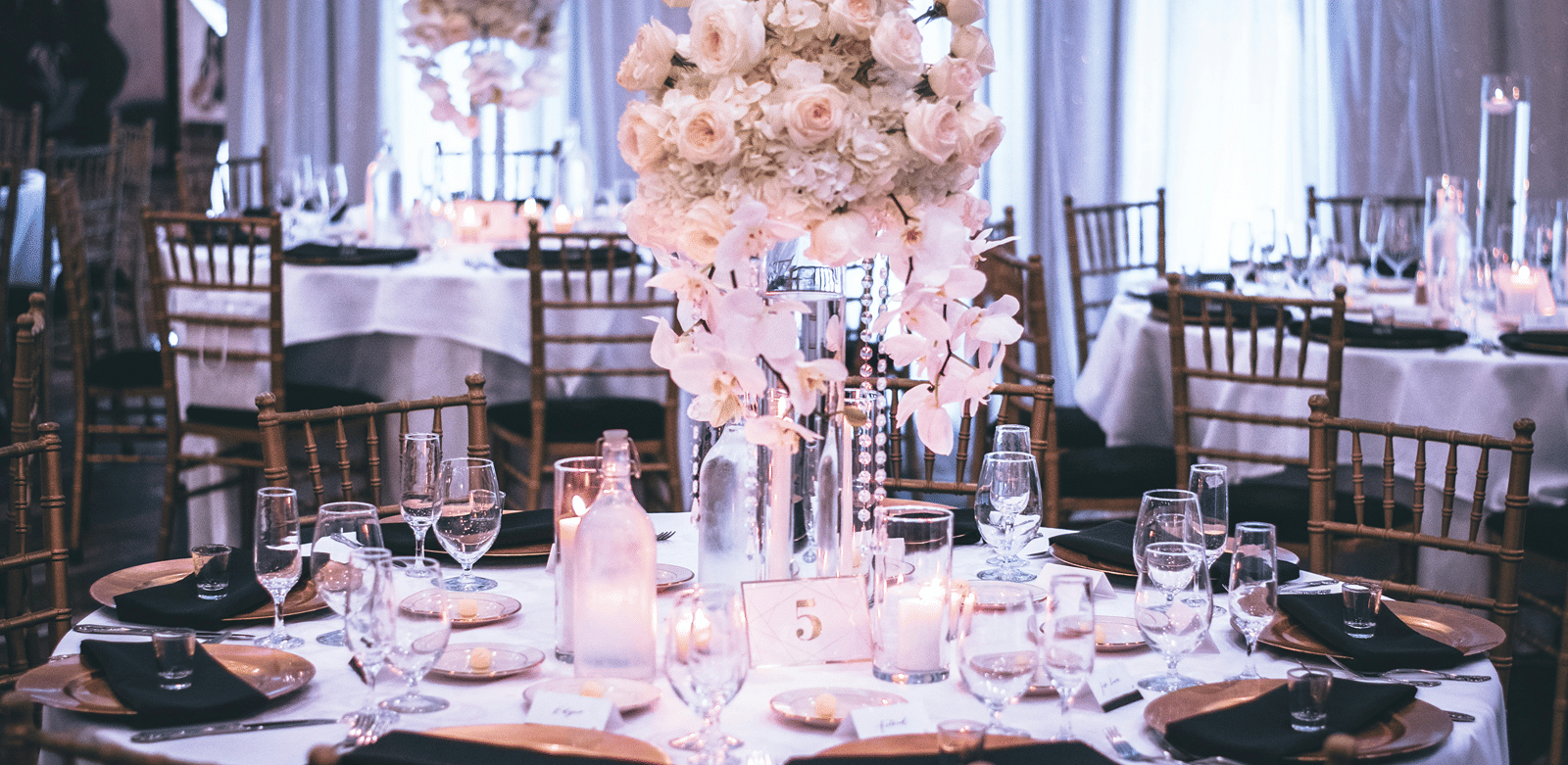 A round table with a white table cloth along with huge plates set on the table and a beautiful flower vase in the middle.