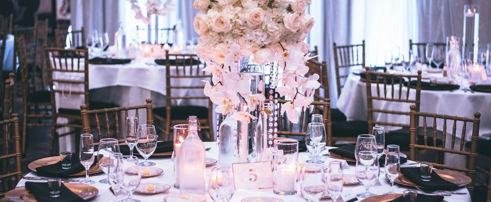 A round table with a white table cloth along with huge plates set on the table and a beautiful flower vase in the middle.