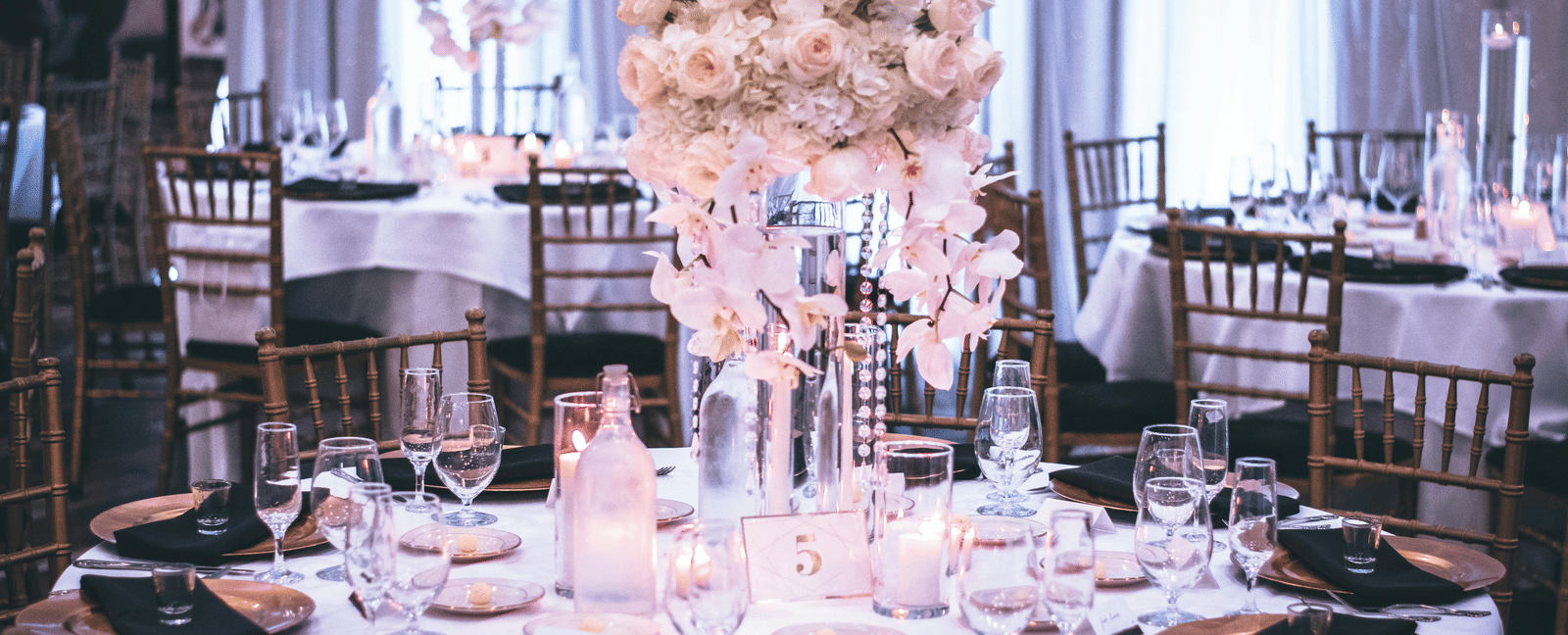 A round table with a white table cloth along with huge plates set on the table and a beautiful flower vase in the middle.